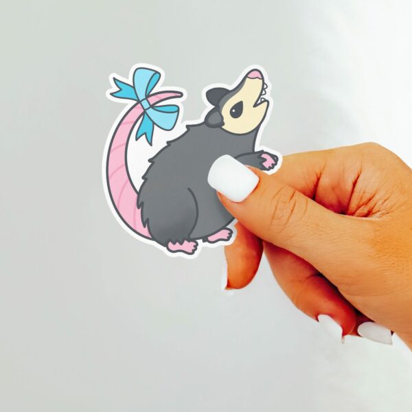 This image shows an hand-drawn adorable sticker, Possum With A Bow Sticker, which is available to purchase from HunnieByte.com