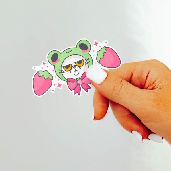 This image shows an hand-drawn adorable sticker, Grumpy Kitty with Frog Hat Sticker, which is available to purchase from HunnieByte.com