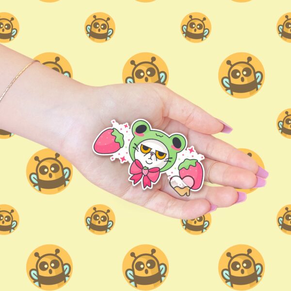 This image shows an hand-drawn adorable sticker, Grumpy Kitty with Frog Hat Sticker, which is available to purchase from HunnieByte.com